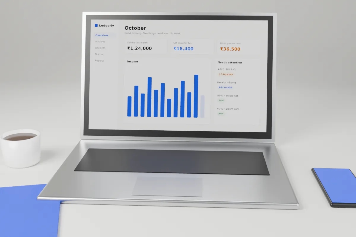 Laptop on a light desk showing the Ledgerly dashboard, with a phone, a notebook and a coffee cup